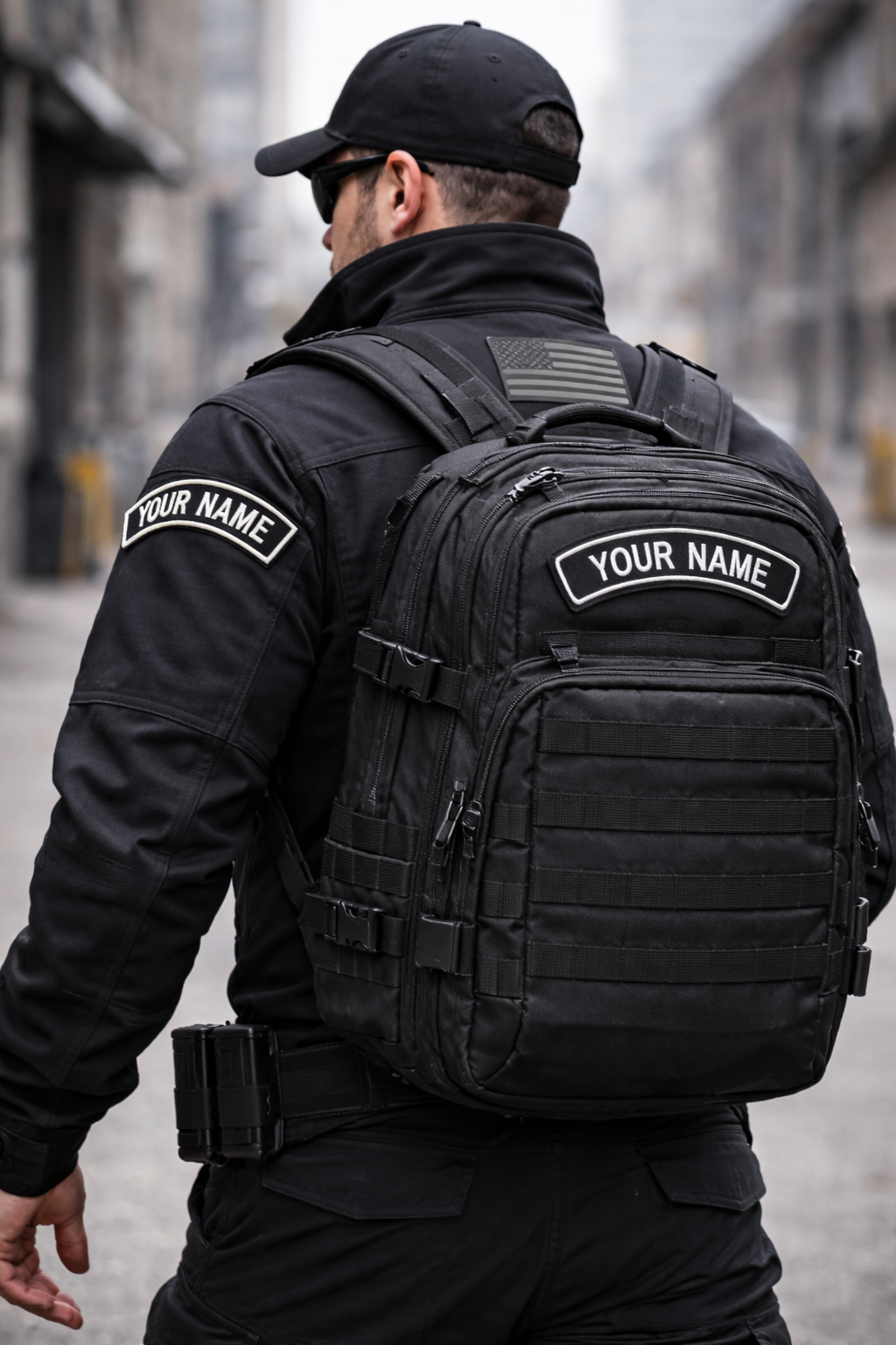 Person wearing a black tactical outfit with a backpack and patches on an urban street.